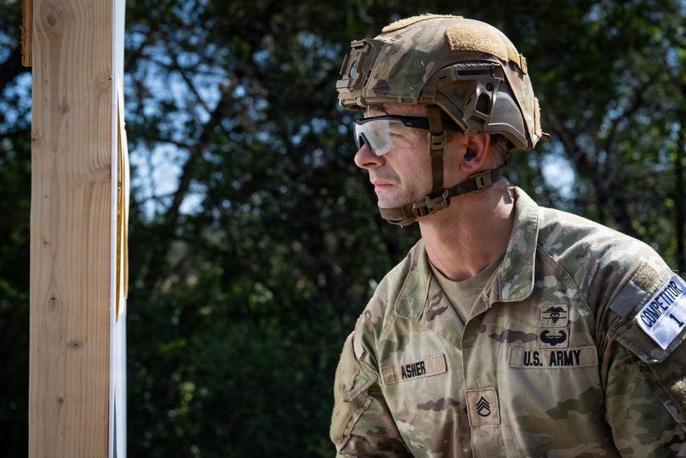 A U.S. Army soldier identified as 'ASHER' by name tape, holding the rank of Staff Sergeant, is photographed in a tactical outdoor setting. He is wearing full combat gear including a multicam uniform, tactical helmet, and protective eyewear. A patch on his right sleeve reads 'COMPETITOR 1', suggesting participation in a military competition or evaluation event.