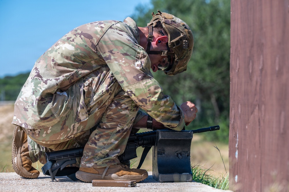 A soldier in full OCP (Operational Camouflage Pattern) uniform is crouched near a wall or barrier, appearing to interact with a dark metal container or ammunition box. The soldier is wearing a tactical helmet with eye protection and tan combat boots. The setting appears to be an outdoor training range or field exercise environment.