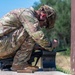 A soldier in full OCP (Operational Camouflage Pattern) uniform is crouched near a wall or barrier, appearing to interact with a dark metal container or ammunition box. The soldier is wearing a tactical helmet with eye protection and tan combat boots. The setting appears to be an outdoor training range or field exercise environment.