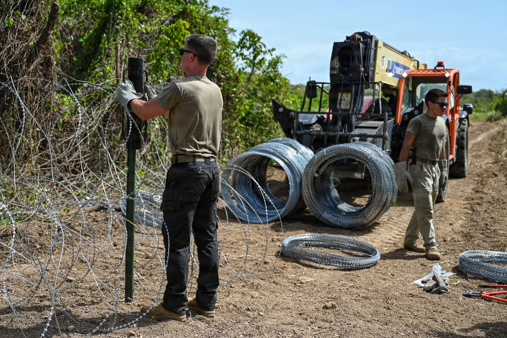 Two individuals in military-style olive drab uniforms and tactical pants are installing concertina (razor) wire along a dirt path or road. One person is securing wire to a green metal post while the other stands near coiled wire rolls and a yellow-and-orange telescopic forklift/telehandler. Multiple coils of concertina wire are staged on the ground and on the equipment nearby.