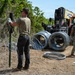 Two individuals in military-style olive drab uniforms and tactical pants are installing concertina (razor) wire along a dirt path or road. One person is securing wire to a green metal post while the other stands near coiled wire rolls and a yellow-and-orange telescopic forklift/telehandler. Multiple coils of concertina wire are staged on the ground and on the equipment nearby.