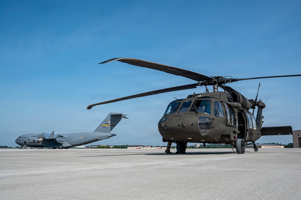 A U.S. Army UH-60 Black Hawk helicopter is parked on a concrete flight line apron, painted in olive drab green. In the background, a U.S. Air Force C-17 Globemaster III transport aircraft is visible on the same tarmac. The image appears to be taken at a joint military airfield under clear blue skies.