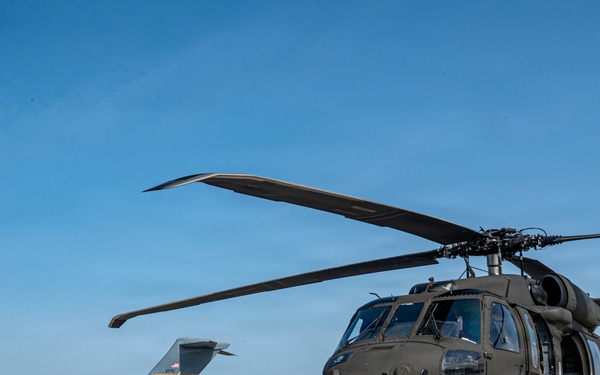 A U.S. Army UH-60 Black Hawk helicopter is parked on a concrete flight line apron, painted in olive drab green. In the background, a U.S. Air Force C-17 Globemaster III transport aircraft is visible on the same tarmac. The image appears to be taken at a joint military airfield under clear blue skies.