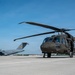 A U.S. Army UH-60 Black Hawk helicopter is parked on a concrete flight line apron, painted in olive drab green. In the background, a U.S. Air Force C-17 Globemaster III transport aircraft is visible on the same tarmac. The image appears to be taken at a joint military airfield under clear blue skies.