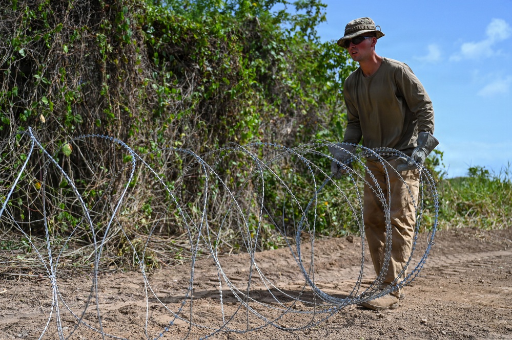 A person in military-style clothing is deploying concertina (razor) wire along a dirt path or perimeter. The individual is wearing tan/olive drab attire consistent with military or security personnel, and is actively unrolling coiled razor wire on the ground. The setting appears to be a rural or semi-arid outdoor environment with dense vegetation in the background.
