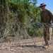 A person in military-style clothing is deploying concertina (razor) wire along a dirt path or perimeter. The individual is wearing tan/olive drab attire consistent with military or security personnel, and is actively unrolling coiled razor wire on the ground. The setting appears to be a rural or semi-arid outdoor environment with dense vegetation in the background.