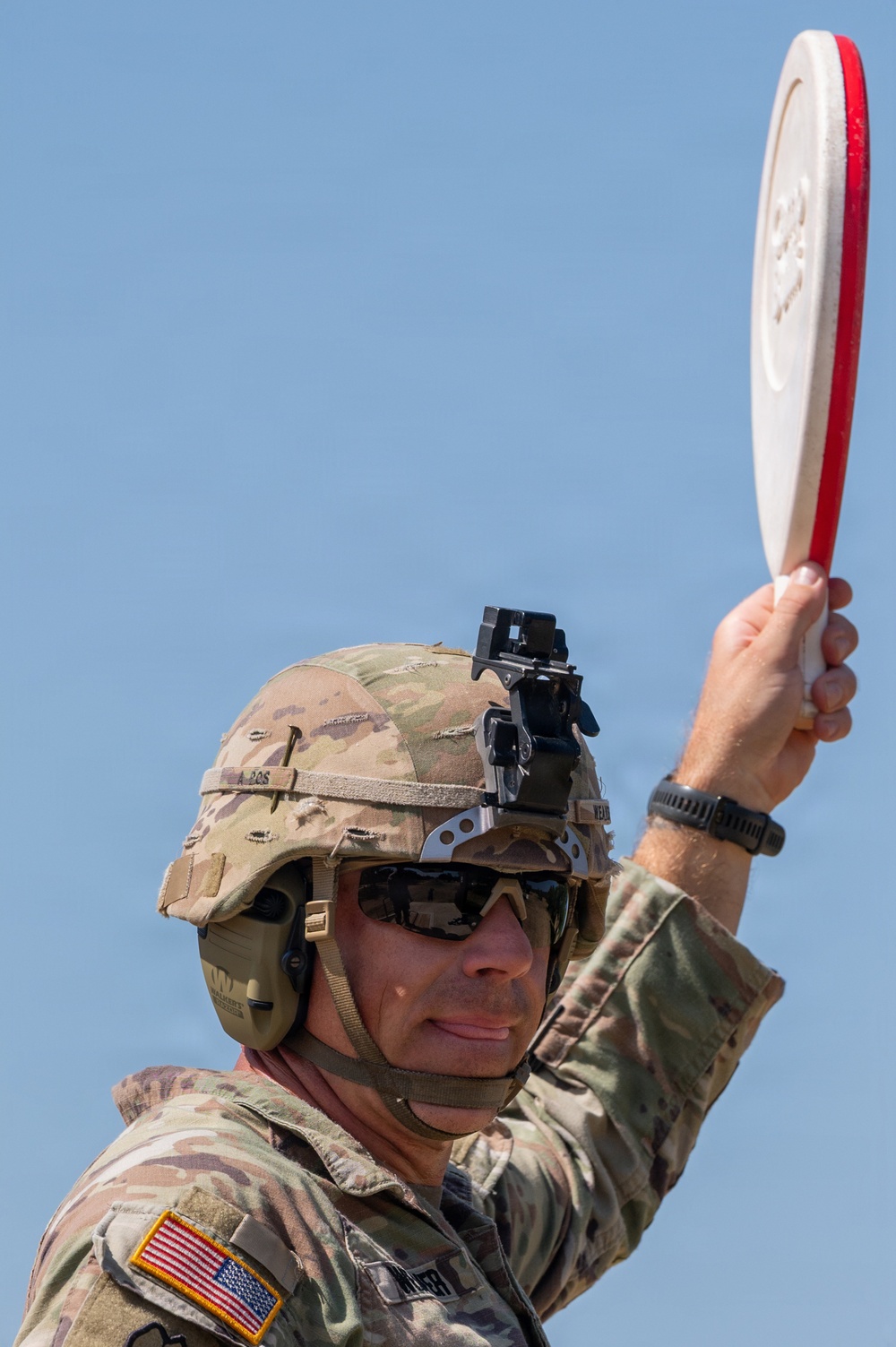 A U.S. Army soldier in full combat gear, including an OCP (Operational Camouflage Pattern) uniform and helmet with night vision mount, holds up a white and red clay pigeon target disk overhead. The soldier is wearing tactical eye protection, hearing protection, and a smartwatch, suggesting participation in a marksmanship or shooting range activity. The American flag patch is visible on the left sleeve, identifying this as a U.S. military personnel.