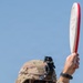 A U.S. Army soldier in full combat gear, including an OCP (Operational Camouflage Pattern) uniform and helmet with night vision mount, holds up a white and red clay pigeon target disk overhead. The soldier is wearing tactical eye protection, hearing protection, and a smartwatch, suggesting participation in a marksmanship or shooting range activity. The American flag patch is visible on the left sleeve, identifying this as a U.S. military personnel.