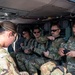 A group of U.S. Air Force personnel in OCP (Operational Camouflage Pattern) uniforms are seated inside a military helicopter, appearing to prepare for or complete a flight. Several personnel are adjusting harnesses and seatbelts, with one female service member standing near the open door. Name tapes and U.S. Air Force branch tapes are visible on multiple individuals.