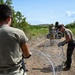Military or security personnel are installing concertina (razor) wire along a dirt path in a tropical or subtropical outdoor environment. At least three individuals in olive/tan military-style uniforms are working together to deploy and position coiled razor wire barriers. The setting appears to be a rural area with dense vegetation and a clear blue sky.