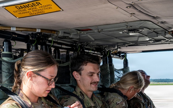 Several U.S. Air Force personnel in OCP (Operational Camouflage Pattern) uniforms are seated inside what appears to be a military helicopter or aircraft cabin. A warning sign reading 'DANGER - EXTREME NOISE HAZARD - HEARING PROTECTION REQUIRED' is visible on the overhead panel. The personnel appear to be preparing for or waiting during a flight operation on an airfield.
