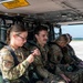Several U.S. Air Force personnel in OCP (Operational Camouflage Pattern) uniforms are seated inside what appears to be a military helicopter or aircraft cabin. A warning sign reading 'DANGER - EXTREME NOISE HAZARD - HEARING PROTECTION REQUIRED' is visible on the overhead panel. The personnel appear to be preparing for or waiting during a flight operation on an airfield.