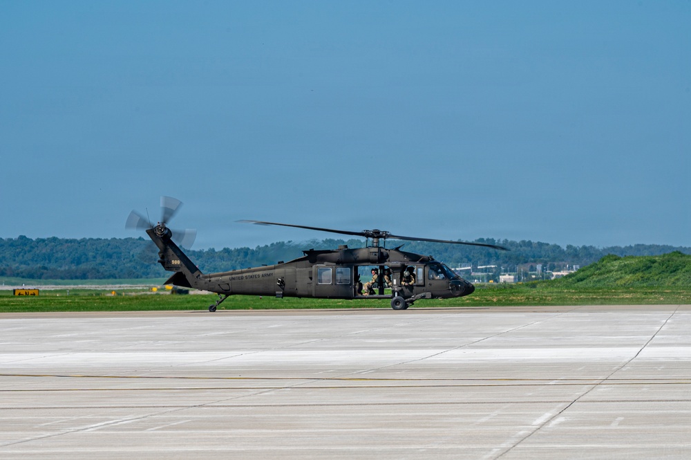 A United States Army UH-60 Black Hawk helicopter is taxiing or hovering just above a concrete airfield tarmac. The aircraft is painted in standard olive drab/dark green military livery with 'UNITED STATES ARMY' markings visible on the fuselage. Crew members are visible through the cockpit windows, and the main rotor blades are in motion.