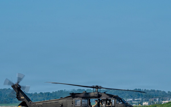 A United States Army UH-60 Black Hawk helicopter is taxiing or hovering just above a concrete airfield tarmac. The aircraft is painted in standard olive drab/dark green military livery with 'UNITED STATES ARMY' markings visible on the fuselage. Crew members are visible through the cockpit windows, and the main rotor blades are in motion.