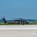 A United States Army UH-60 Black Hawk helicopter is taxiing or hovering just above a concrete airfield tarmac. The aircraft is painted in standard olive drab/dark green military livery with 'UNITED STATES ARMY' markings visible on the fuselage. Crew members are visible through the cockpit windows, and the main rotor blades are in motion.