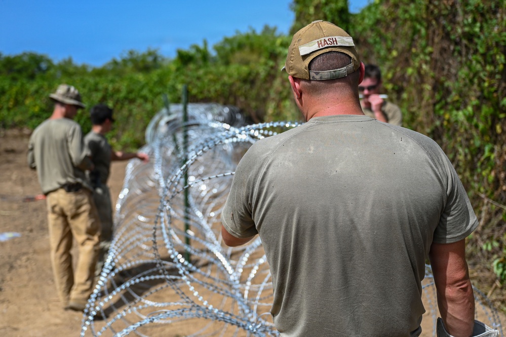 Several workers in military-style or utility clothing are installing concertina (razor) wire along a dirt path bordered by dense vegetation. The foreground figure wears a cap labeled 'HASH' and a soiled olive t-shirt, actively handling the coiled wire. The scene appears to be a perimeter security or barrier installation operation in an outdoor, tropical-looking environment.