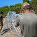 Several workers in military-style or utility clothing are installing concertina (razor) wire along a dirt path bordered by dense vegetation. The foreground figure wears a cap labeled 'HASH' and a soiled olive t-shirt, actively handling the coiled wire. The scene appears to be a perimeter security or barrier installation operation in an outdoor, tropical-looking environment.