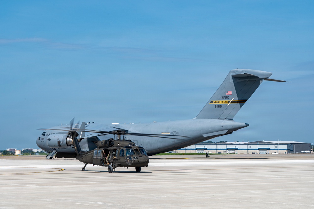 A U.S. Air Force C-17 Globemaster III cargo aircraft assigned to the Air Force Reserve Command (AFRC) Pittsburgh unit, tail number 91189, is parked on a tarmac. In the foreground, a U.S. Army UH-60 Black Hawk helicopter is positioned directly in front of the large transport aircraft. The scene appears to be at a military airfield with hangars visible in the background.
