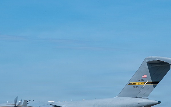 A U.S. Air Force C-17 Globemaster III cargo aircraft assigned to the Air Force Reserve Command (AFRC) Pittsburgh unit, tail number 91189, is parked on a tarmac. In the foreground, a U.S. Army UH-60 Black Hawk helicopter is positioned directly in front of the large transport aircraft. The scene appears to be at a military airfield with hangars visible in the background.