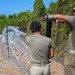 Two individuals in military-style olive/tan uniforms are installing concertina (razor) wire along a dirt path bordered by dense vegetation. One person is driving a metal post into the ground while the other assists with positioning. The scene appears to be a border security or perimeter fortification operation conducted in an outdoor, rural environment.