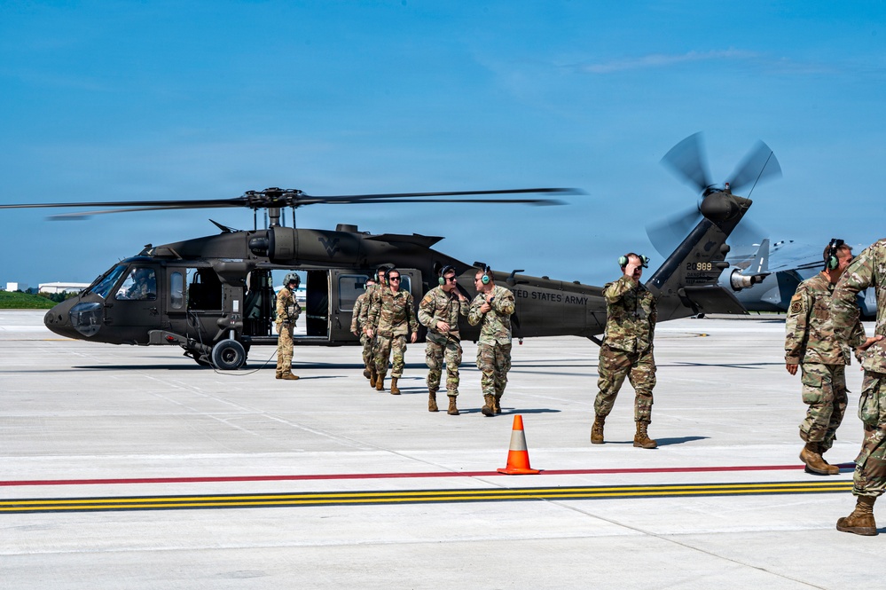 A group of U.S. Army soldiers are gathered on a military airfield tarmac near a black UH-60 Black Hawk helicopter with its rotors spinning. The personnel are wearing OCP (Operational Camouflage Pattern) uniforms and hearing protection, suggesting active flight operations. The scene appears to depict a post-landing or pre-departure briefing on the flight line.