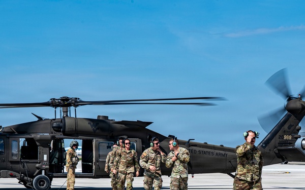 A group of U.S. Army soldiers are gathered on a military airfield tarmac near a black UH-60 Black Hawk helicopter with its rotors spinning. The personnel are wearing OCP (Operational Camouflage Pattern) uniforms and hearing protection, suggesting active flight operations. The scene appears to depict a post-landing or pre-departure briefing on the flight line.