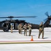 A group of U.S. Army soldiers are gathered on a military airfield tarmac near a black UH-60 Black Hawk helicopter with its rotors spinning. The personnel are wearing OCP (Operational Camouflage Pattern) uniforms and hearing protection, suggesting active flight operations. The scene appears to depict a post-landing or pre-departure briefing on the flight line.