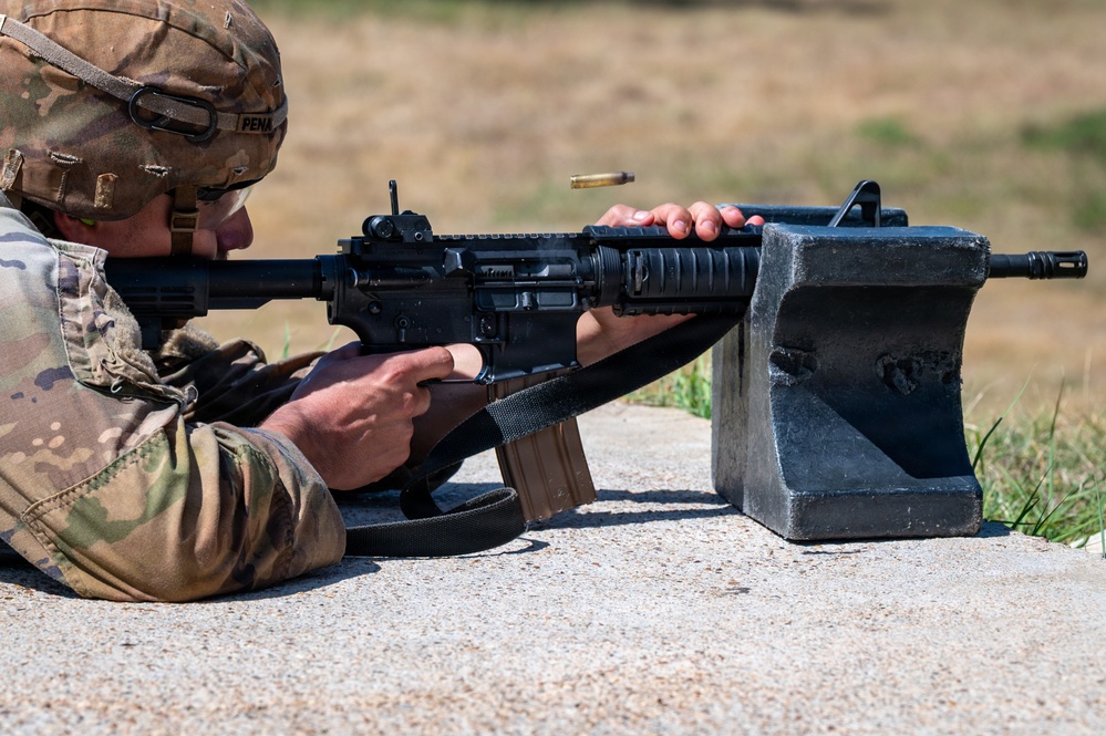 A soldier in MultiCam/OCP camouflage uniform is firing an M4/M16-style rifle from a prone position on a concrete surface at what appears to be a military firing range. A spent brass cartridge casing is visibly ejecting from the weapon's ejection port, captured mid-flight. The soldier's name tape reads 'PENA' and the rifle is rested on a concrete shooting block.