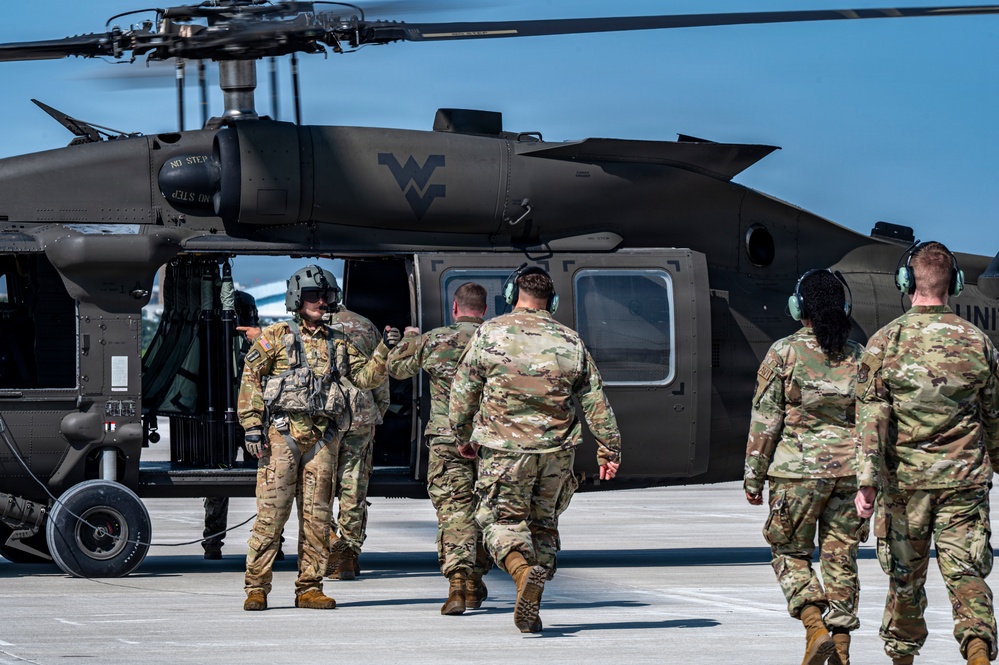Military personnel in OCP (Operational Camouflage Pattern) uniforms approach a dark olive/black UH-60 Black Hawk helicopter on a tarmac. The helicopter bears the West Virginia National Guard 'Flying WV' logo on its fuselage. A crew member wearing a flight helmet appears to be directing or briefing the approaching soldiers.