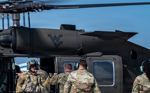 Military personnel in OCP (Operational Camouflage Pattern) uniforms approach a dark olive/black UH-60 Black Hawk helicopter on a tarmac. The helicopter bears the West Virginia National Guard 'Flying WV' logo on its fuselage. A crew member wearing a flight helmet appears to be directing or briefing the approaching soldiers.