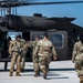 Military personnel in OCP (Operational Camouflage Pattern) uniforms approach a dark olive/black UH-60 Black Hawk helicopter on a tarmac. The helicopter bears the West Virginia National Guard 'Flying WV' logo on its fuselage. A crew member wearing a flight helmet appears to be directing or briefing the approaching soldiers.