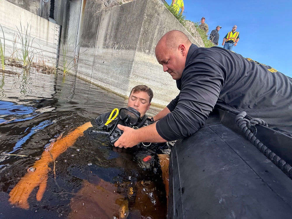 A diver in full scuba gear is in dark water near a concrete structure while a bald man in a black long-sleeve shirt assists him from what appears to be an inflatable boat. Several observers wearing high-visibility vests and military-style clothing are visible on an elevated area in the background. The scene appears to be a dive operation or training exercise near a water control structure or lock.