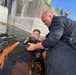A diver in full scuba gear is in dark water near a concrete structure while a bald man in a black long-sleeve shirt assists him from what appears to be an inflatable boat. Several observers wearing high-visibility vests and military-style clothing are visible on an elevated area in the background. The scene appears to be a dive operation or training exercise near a water control structure or lock.