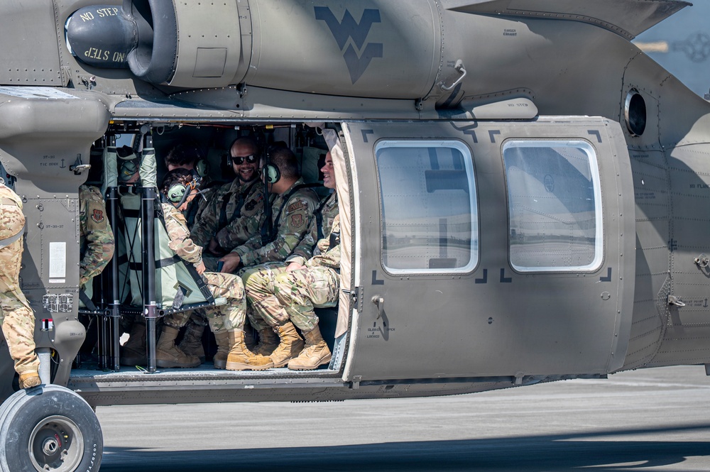 Military personnel in camouflage uniforms are seated inside the open cabin of a military helicopter, likely a UH-60 Black Hawk, on a tarmac or airfield. Several soldiers appear relaxed and smiling, suggesting a pre- or post-mission moment. A West Virginia National Guard insignia is visible on the fuselage.