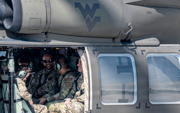 Military personnel in camouflage uniforms are seated inside the open cabin of a military helicopter, likely a UH-60 Black Hawk, on a tarmac or airfield. Several soldiers appear relaxed and smiling, suggesting a pre- or post-mission moment. A West Virginia National Guard insignia is visible on the fuselage.