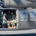 Military personnel in camouflage uniforms are seated inside the open cabin of a military helicopter, likely a UH-60 Black Hawk, on a tarmac or airfield. Several soldiers appear relaxed and smiling, suggesting a pre- or post-mission moment. A West Virginia National Guard insignia is visible on the fuselage.