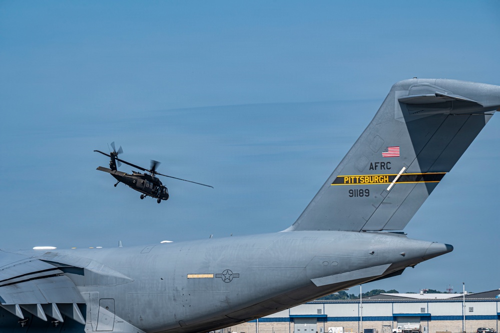 A U.S. Army Black Hawk helicopter flies in the background while the tail section of a U.S. Air Force Reserve Command (AFRC) C-17 Globemaster III, tail number 91189, assigned to Pittsburgh, is prominently visible in the foreground. The scene appears to be at Pittsburgh International Airport or an associated Air Reserve Station. Both aircraft are military assets operating in the same airspace.