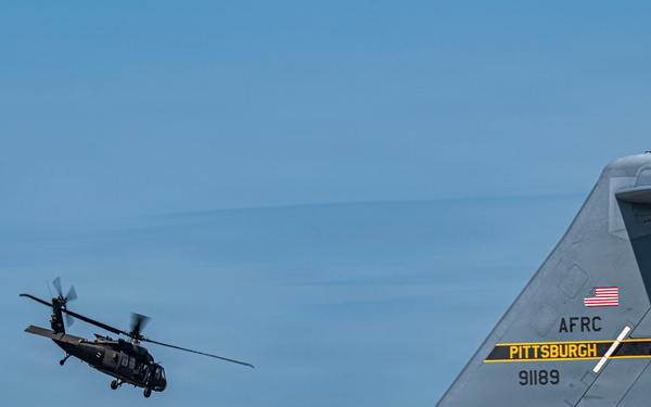 A U.S. Army Black Hawk helicopter flies in the background while the tail section of a U.S. Air Force Reserve Command (AFRC) C-17 Globemaster III, tail number 91189, assigned to Pittsburgh, is prominently visible in the foreground. The scene appears to be at Pittsburgh International Airport or an associated Air Reserve Station. Both aircraft are military assets operating in the same airspace.