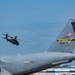 A U.S. Army Black Hawk helicopter flies in the background while the tail section of a U.S. Air Force Reserve Command (AFRC) C-17 Globemaster III, tail number 91189, assigned to Pittsburgh, is prominently visible in the foreground. The scene appears to be at Pittsburgh International Airport or an associated Air Reserve Station. Both aircraft are military assets operating in the same airspace.