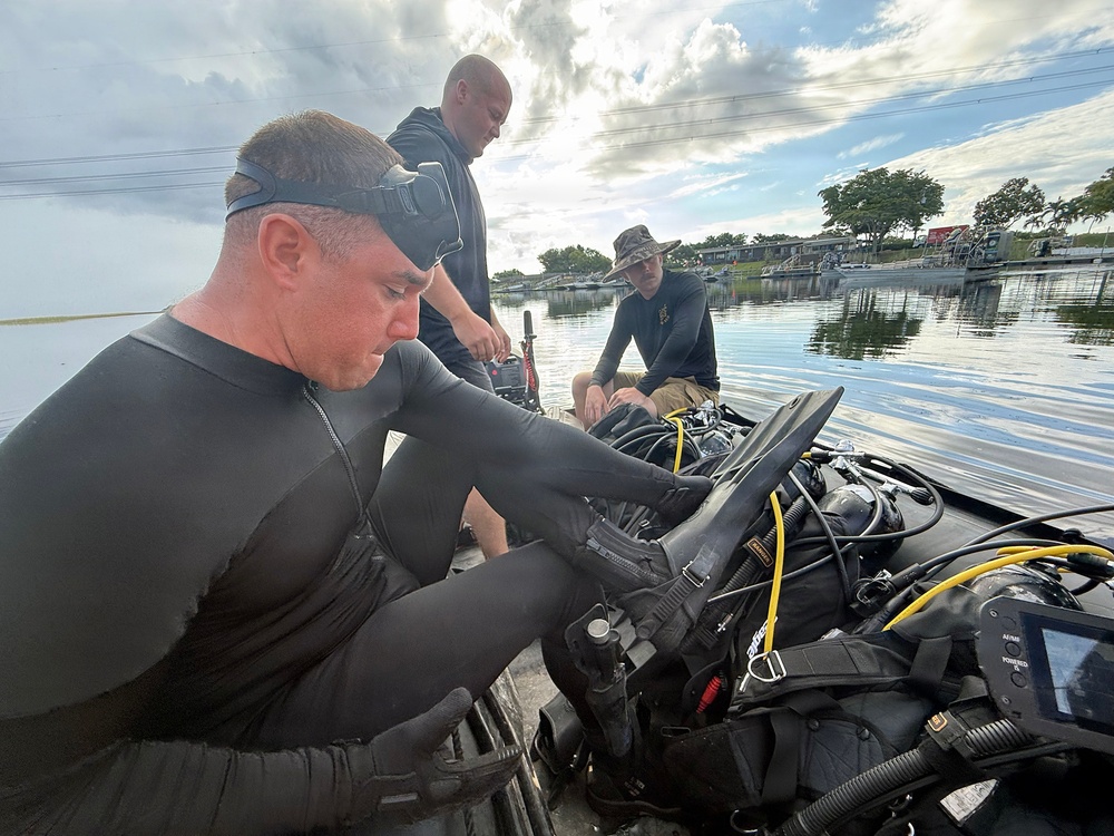Three individuals are on a small boat on a calm waterway, preparing diving equipment. The person in the foreground is wearing a black wetsuit with a dive mask pushed up on his forehead, handling scuba gear. The setting appears to be a flooded or low-lying coastal area, possibly during or after a weather event given the dramatic cloud cover.