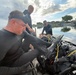 Three individuals are on a small boat on a calm waterway, preparing diving equipment. The person in the foreground is wearing a black wetsuit with a dive mask pushed up on his forehead, handling scuba gear. The setting appears to be a flooded or low-lying coastal area, possibly during or after a weather event given the dramatic cloud cover.