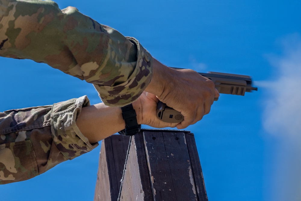 A person wearing military OCP (Operational Camouflage Pattern) uniform is firing a semi-automatic pistol, captured from a low angle against a clear blue sky. The pistol appears to be a SIG Sauer M17/M18 or similar model with a flat dark earth (FDE) finish, consistent with U.S. military issue. Visible propellant gas discharge from the muzzle indicates the weapon has just been fired.