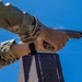 A person wearing military OCP (Operational Camouflage Pattern) uniform is firing a semi-automatic pistol, captured from a low angle against a clear blue sky. The pistol appears to be a SIG Sauer M17/M18 or similar model with a flat dark earth (FDE) finish, consistent with U.S. military issue. Visible propellant gas discharge from the muzzle indicates the weapon has just been fired.