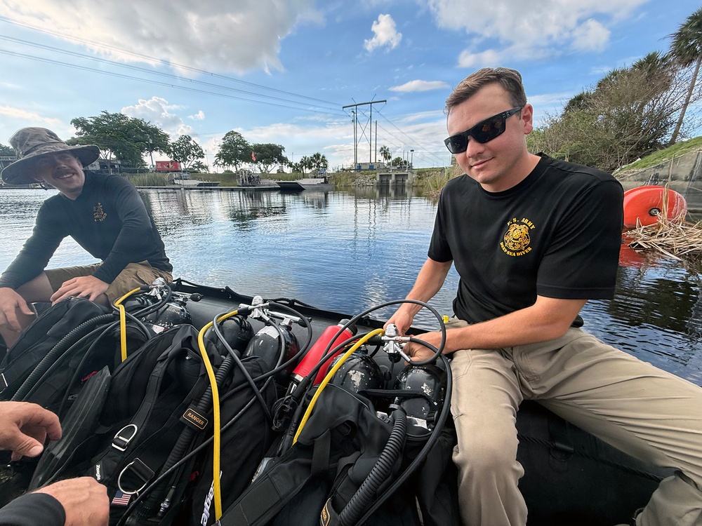 Two individuals are seated on an inflatable boat on a calm waterway, surrounded by scuba diving equipment. The person in the foreground wears a black U.S. Army t-shirt with a logo and sunglasses, and appears to be handling diving gear. The setting appears to be a Florida waterway near a water control structure or lock.