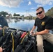 Two individuals are seated on an inflatable boat on a calm waterway, surrounded by scuba diving equipment. The person in the foreground wears a black U.S. Army t-shirt with a logo and sunglasses, and appears to be handling diving gear. The setting appears to be a Florida waterway near a water control structure or lock.