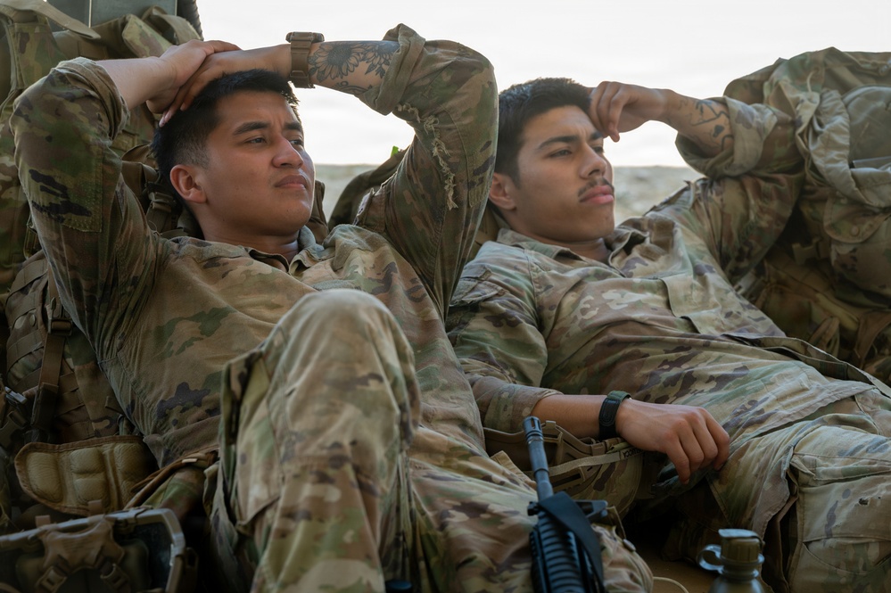 Two soldiers in U.S. Army Operational Camouflage Pattern (OCP) uniforms are resting in a relaxed posture, leaning back with their hands behind or near their heads. Both appear to be taking a break, possibly during a field exercise or deployment. A rifle is visible in the foreground between them.