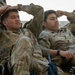 Two soldiers in U.S. Army Operational Camouflage Pattern (OCP) uniforms are resting in a relaxed posture, leaning back with their hands behind or near their heads. Both appear to be taking a break, possibly during a field exercise or deployment. A rifle is visible in the foreground between them.