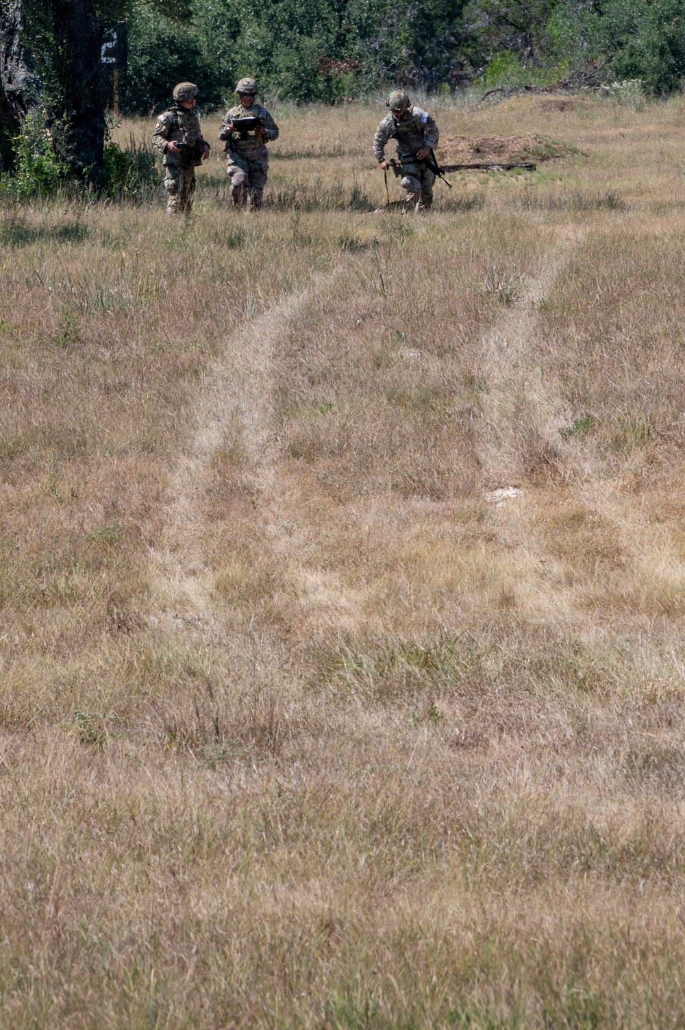 Three military personnel in camouflage uniforms are moving through an open grassy field toward a tree line. One individual appears to be holding a tablet or device, while another carries a rifle. The setting suggests a field training exercise in a dry, open terrain environment.