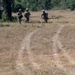 Three military personnel in camouflage uniforms are moving through an open grassy field toward a tree line. One individual appears to be holding a tablet or device, while another carries a rifle. The setting suggests a field training exercise in a dry, open terrain environment.