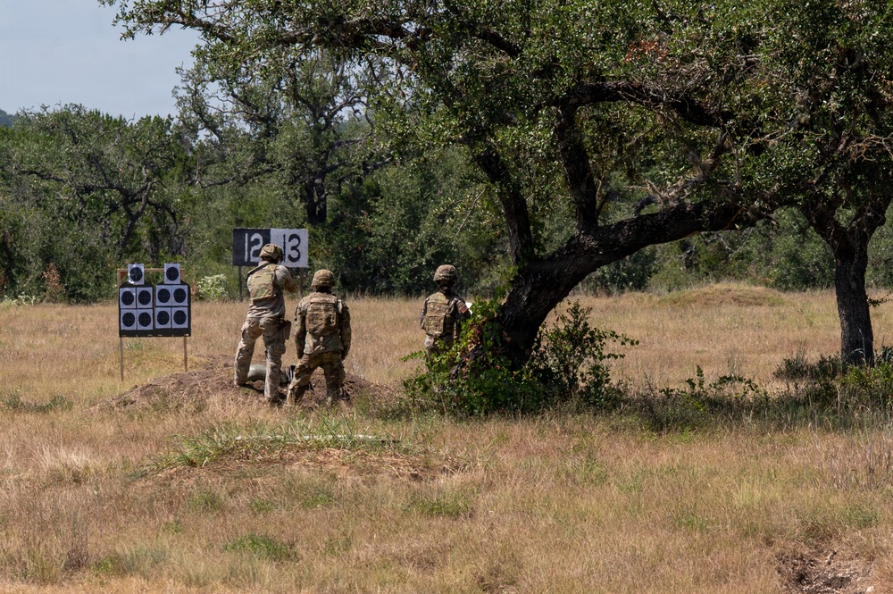 Three military personnel in camouflage uniforms and tactical gear are gathered near shooting range targets in an open field. The targets are numbered 12 and 13, with circular black-and-white scoring targets visible on stands. The setting appears to be an outdoor military training range with dry grassland and oak trees in the background.